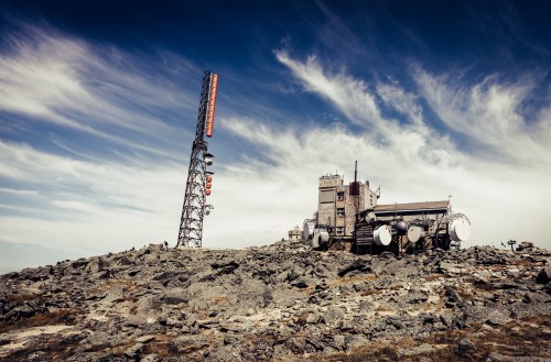 A rocky landscape with a tall communication tower and a building against a backdrop of blue sky and wispy clouds.