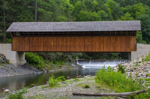 A wooden covered bridge spans a river, with a small waterfall and lush greenery in the background.