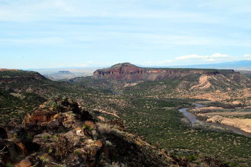 A panoramic view of a rugged landscape with a river winding through green hills and distant mesas under a blue sky.
