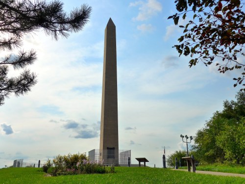 A tall obelisk stands against a cloudy sky, surrounded by greenery and flowers, with informational signs nearby.