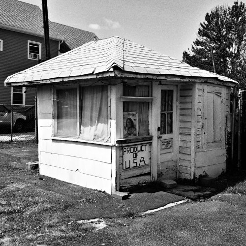 A small, weathered white building with a sign reading "Product of USA," surrounded by grass and nearby structures.