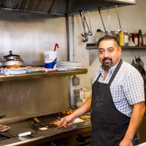 A chef in a checkered shirt and apron stands by a grill in a busy kitchen, preparing food with a smile.