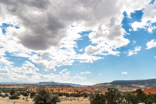 A vast landscape featuring red rock formations, green shrubs, and a cloudy blue sky.