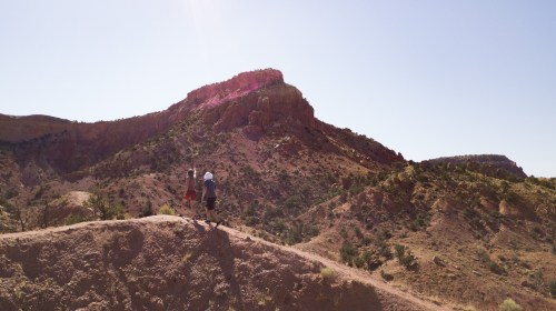 Two hikers walk along a ridge with a rocky mountain in the background under a clear blue sky.