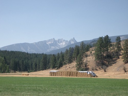 A scenic landscape featuring stacked hay bales, green fields, and mountains in the background under a clear blue sky.