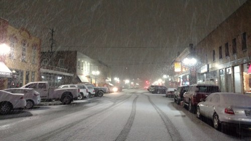 Snowy street scene at night, with falling snow, parked cars, and illuminated storefronts lining the road.
