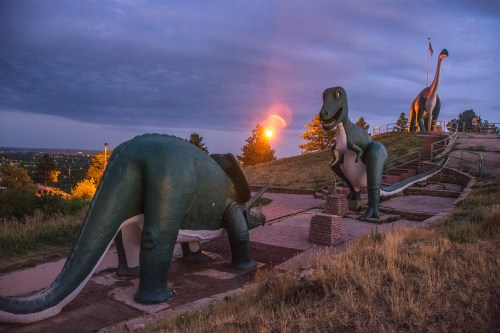 Two large dinosaur statues on a hillside at sunset, with a dramatic sky and distant city lights visible.