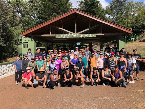 A large group of people poses together in front of a wooden pavilion at Camp Palehua, surrounded by trees.