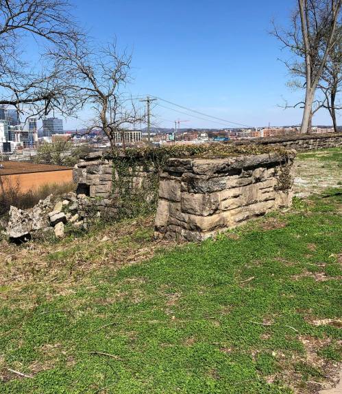 Stone structure on grassy land with a city skyline in the background under a clear blue sky.