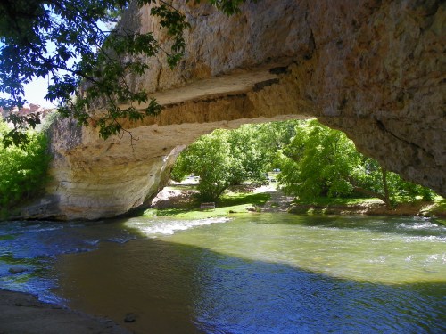 A natural stone arch over a river, surrounded by lush green trees and a sunny blue sky.