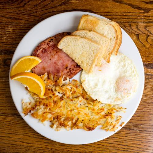 A plate with hash browns, a fried egg, ham, and toast, garnished with orange slices, on a wooden table.