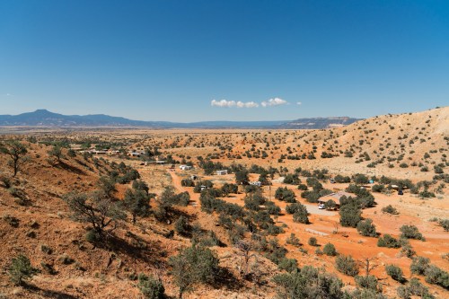 A panoramic view of a desert landscape with red earth, scattered trees, and distant mountains under a clear blue sky.
