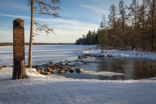A snowy landscape with a river, rocks, and a wooden sign detailing historical information about the area.