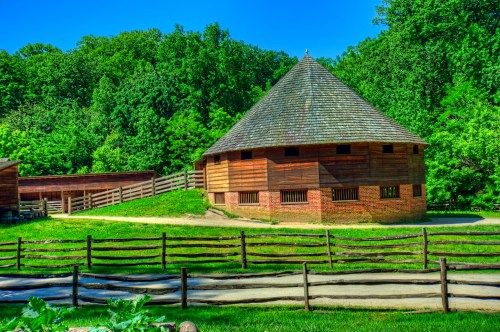 A round wooden building with a conical roof, surrounded by green trees and a wooden fence in a sunny landscape.
