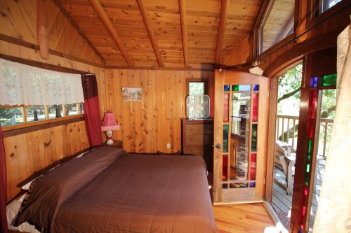 Cozy wooden bedroom with a brown bedspread, colorful stained glass door, and large windows letting in natural light.
