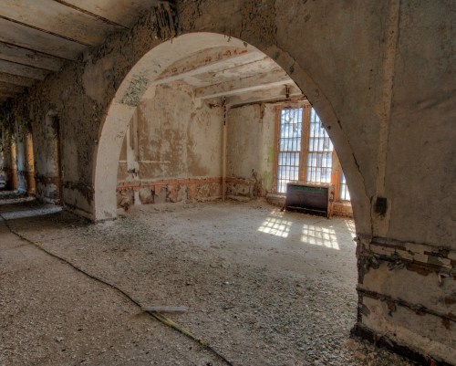 Abandoned room with peeling walls, arched doorway, and sunlight streaming through a window, creating a somber atmosphere.