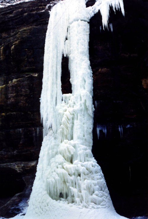 A tall, frozen waterfall with icicles hanging from a rocky cliff, surrounded by a winter landscape.
