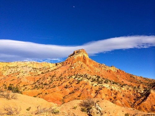 A rugged orange mountain under a clear blue sky with wispy clouds and a small moon in the background.