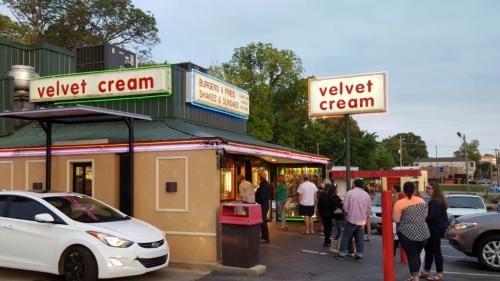 A busy fast-food restaurant named "Velvet Cream" with neon signs, featuring a line of customers outside.