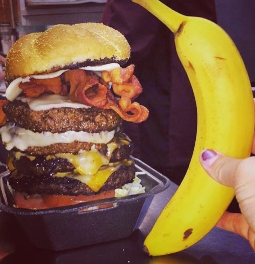 A towering burger with multiple patties, cheese, and bacon, next to a hand holding a banana for scale.