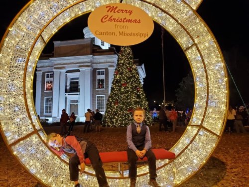 Two boys sit on a festive bench with a Christmas tree and lights in the background, celebrating the holiday season.