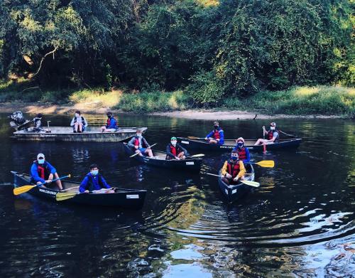 A group of people in canoes and a kayak on a calm river, wearing life jackets and enjoying a sunny day outdoors.