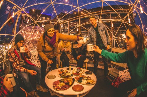 A group of friends enjoying food and drinks inside a cozy, illuminated dome, surrounded by festive lights.