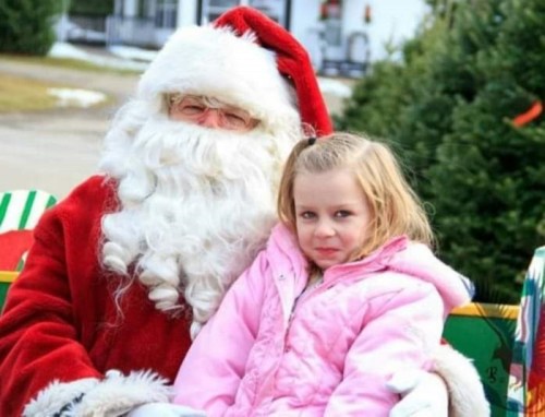 A young girl in a pink coat sits on Santa's lap, smiling, with Christmas trees in the background.