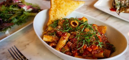 A bowl of rigatoni pasta topped with marinara sauce, herbs, and a slice of garlic bread on the side.