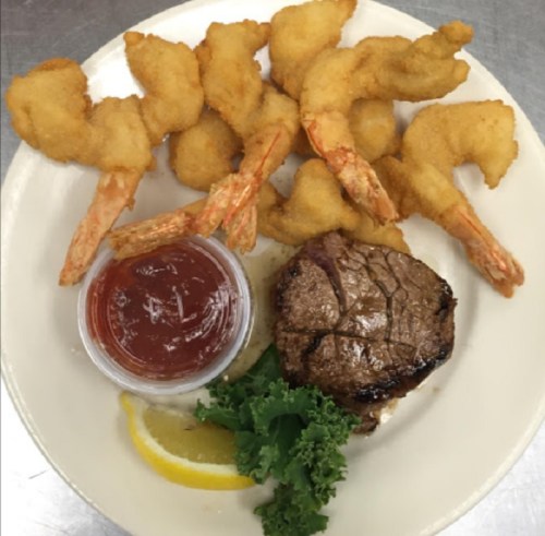 A plate featuring fried shrimp, a grilled steak, a lemon wedge, and a small cup of cocktail sauce.