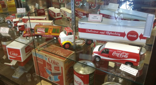 Display of vintage Coca-Cola toy trucks and memorabilia in a glass case.