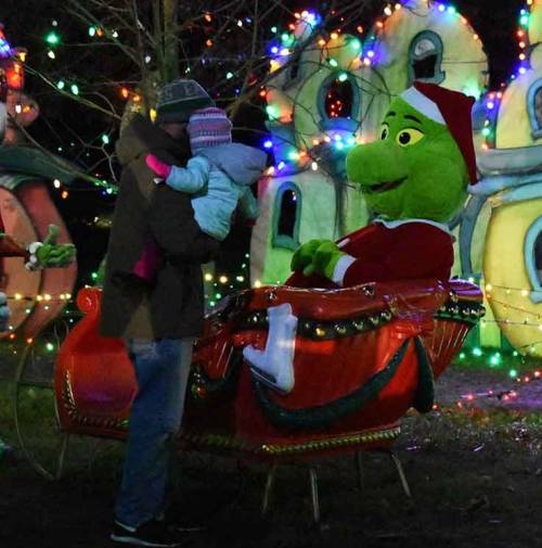 A person holds a child near a festive Grinch figure in a decorated holiday setting with colorful lights.