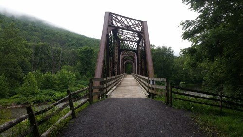 A metal bridge spans a river, surrounded by lush greenery and misty mountains in the background.