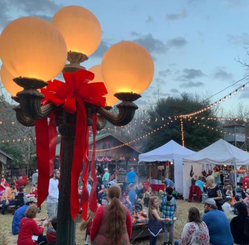 A festive outdoor gathering with people around a fire, decorated with lights and a lamp adorned with a red bow.