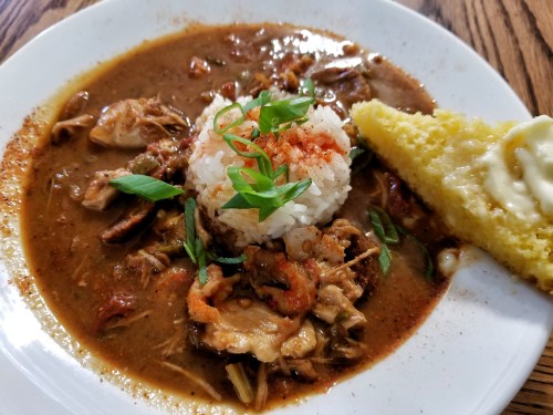 A bowl of gumbo with shrimp, chicken, and rice, served with a slice of cornbread on the side.