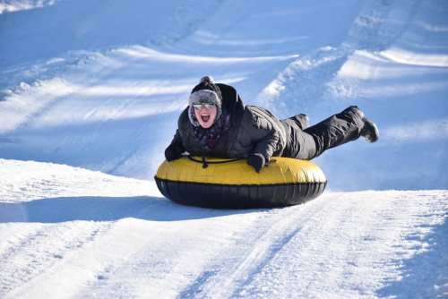 A person in winter gear joyfully sleds down a snowy hill on a yellow and black tube.
