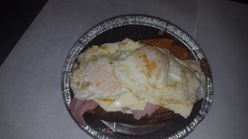 A dish featuring a toasted bun topped with ham, melted cheese, and two fried eggs in a foil container.