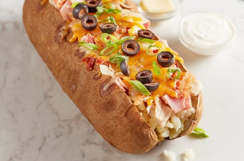 A loaded baked potato topped with cheese, ham, olives, green onions, and served with sour cream on the side.