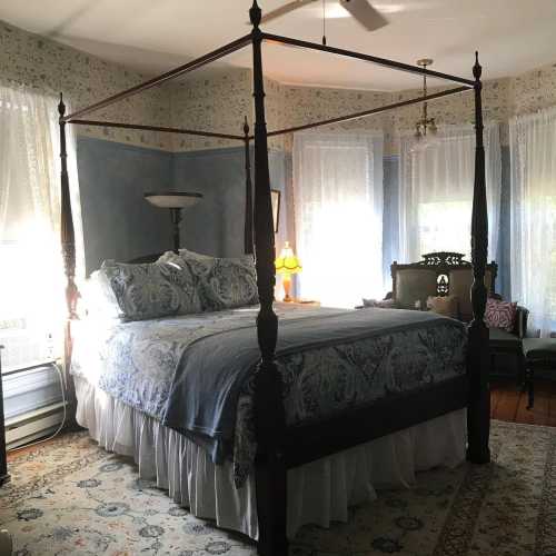 A cozy bedroom featuring a four-poster bed, patterned wallpaper, and large windows with sheer curtains.