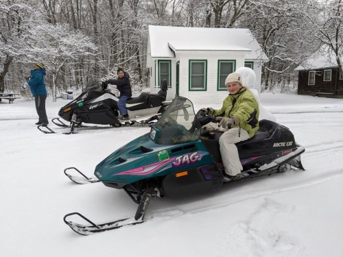Two people on snowmobiles in a snowy landscape, with a small cabin in the background. One person is seated on a snowmobile.