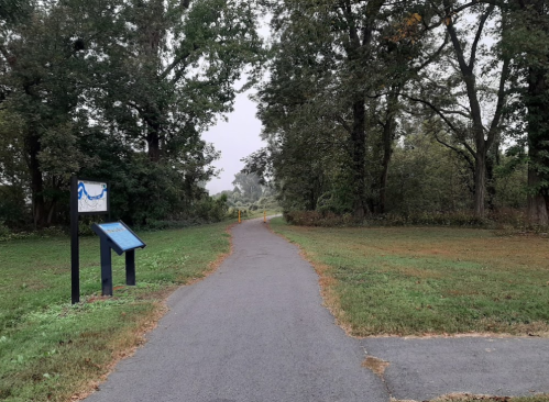 A paved path through a grassy area, flanked by trees, with an informational sign on the left. Overcast sky above.