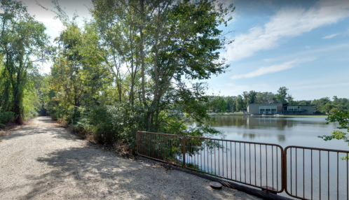 A serene lakeside path lined with trees, leading to a modern building across the water under a clear blue sky.