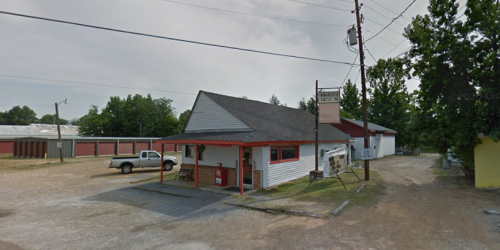 A small, single-story building with a red and white exterior, surrounded by gravel and trees, with a truck parked outside.
