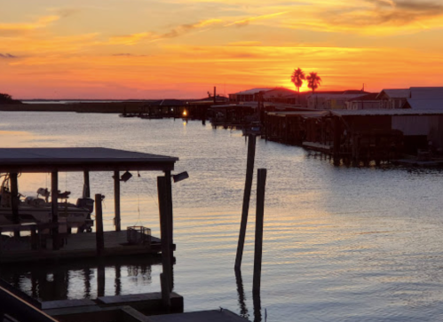Sunset over a calm waterway, with silhouettes of boats and docks against a vibrant orange and purple sky.