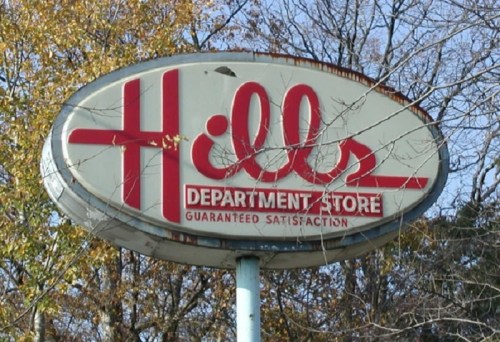 Sign for Hills Department Store, featuring bold red lettering and the tagline "Guaranteed Satisfaction," surrounded by trees.