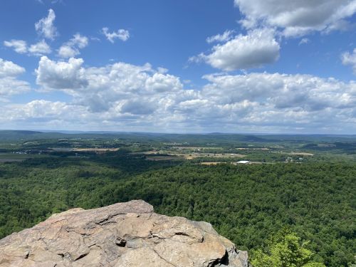 A panoramic view from a rocky overlook, showcasing lush green hills and a blue sky dotted with fluffy clouds.