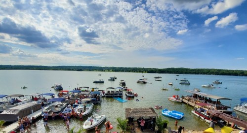 A busy lakeside scene with boats docked, people swimming, and a cloudy sky above.