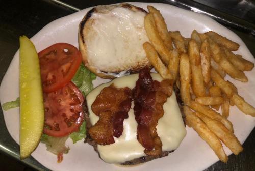 A plate with a cheeseburger topped with bacon, lettuce, and tomato, served with fries and a pickle on the side.