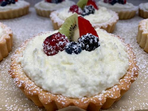 Close-up of creamy fruit tarts topped with berries and kiwi, dusted with powdered sugar on a light background.