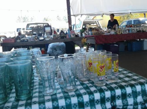 A table with various glassware and collectibles at a market, with a vendor in the background under a tent.
